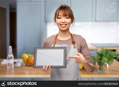 cooking, technology and people concept - happy smiling female chef or waitress in apron showing tablet pc computer with empty screen over home kitchen background. happy woman in apron with tablet pc in kitchen