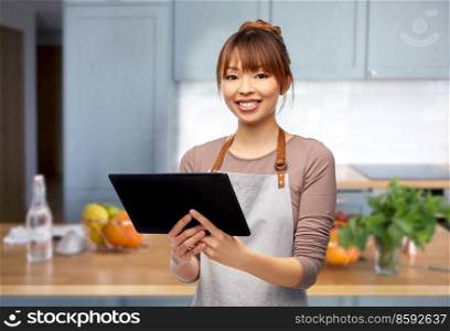 cooking, technology and people concept - happy smiling female chef or waitress in apron with tablet pc computer over home kitchen background. happy woman in apron with tablet pc in kitchen