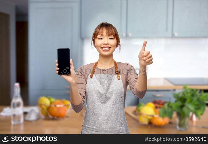 cooking, technology and people concept - happy smiling female chef or waitress in apron showing smartphone with empty screen over home kitchen background. happy woman in apron showing smartphone in kitchen