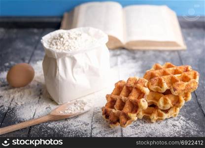 Cooking theme image with a bunch of delicious Belgian waffles, egg, a bag of flour and an open cookbook, on a rustic wooden table.
