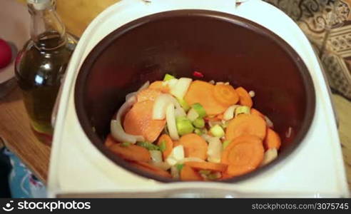 Cooking woman adding chopped tomatoes with knife from wooden cutting board into multicooker and stirring slowly ingredients with spatula