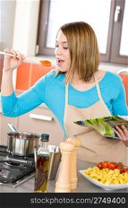 Cooking - Young woman tasting Italian tomato sauce in modern kitchen, with tomatoes and tortellini pasta