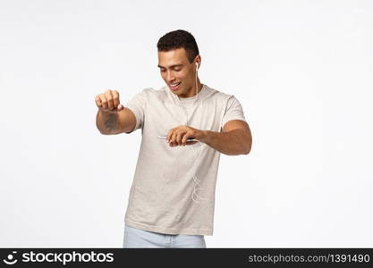 Cool, sexy young handsome man in casual t-shirt, dancing carefree, close eyes and smiling pleased, listen music in wired earphones, lift hands moving to rhythm, enjoy favorite song, hold smartphone.. Cool, sexy young handsome man in casual t-shirt, dancing carefree, close eyes and smiling pleased, listen music in wired earphones, lift hands moving to rhythm, enjoy favorite song, hold smartphone