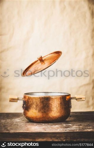 Copper cooking pot with flying open lid on wooden kitchen table at wall background, front view