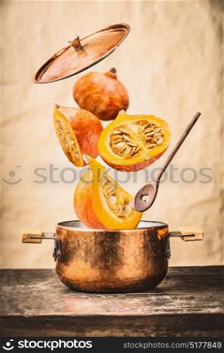 Copper cooking pot with flying pumpkin with knife , open lid and wooden spoon on table, at wall background , front view. Autumn seasonal recipes and eating concept