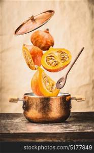 Copper cooking pot with flying pumpkin with knife , open lid and wooden spoon on table, at wall background , front view. Autumn seasonal recipes and eating concept