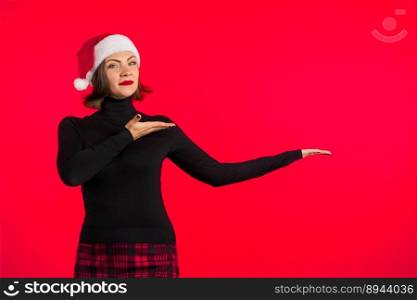 Copy space. Trendy hipster girl in black hat and plaid mini skirt showing to her left side. Smiling lady. Portrait of young pretty woman on red background in studio. High quality photo. Copy space. Pretty woman in black hat plaid mini skirt showing to her left side