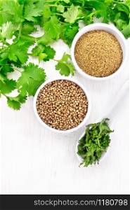 Coriander seeds and ground in two bowls, dried cilantro in a spoon, seasoning greens on wooden board background from above