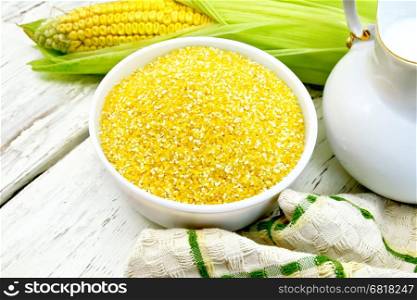 Corn grits in a white bowl, cobs, a jug of milk and a napkin on the background light wooden boards