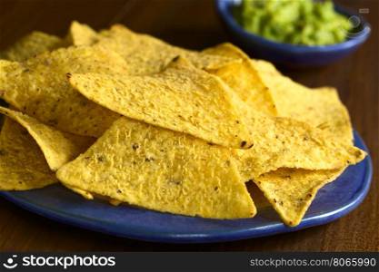 Corn tortilla chips with avocado dip in the back, photographed with natural light (Selective Focus, Focus on the front edge of the tortilla chip in the middle of the image)