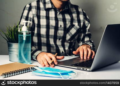 Coronavirus disease or COVID-19 protection concept - Young man work in office room at home with protective and cleaning equipment to protect against corona virus while using laptop computer at desk.