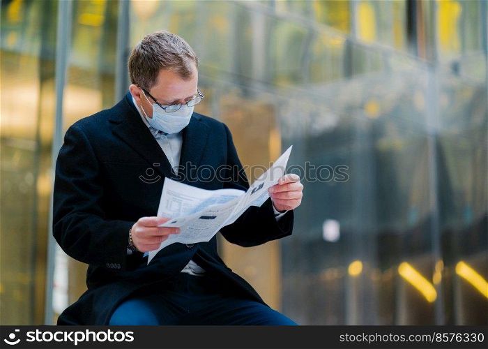 Coronavirus epidemic in city. Horizontal shot of serious man reads newspaper attentively, poses against blurred building background, wears mask for spreading of coronavirus. Businessman with press