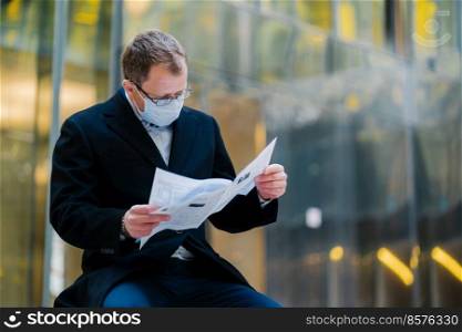 Coronavirus epidemic in city. Horizontal shot of serious man reads newspaper attentively, poses against blurred building background, wears mask for spreading of coronavirus. Businessman with press