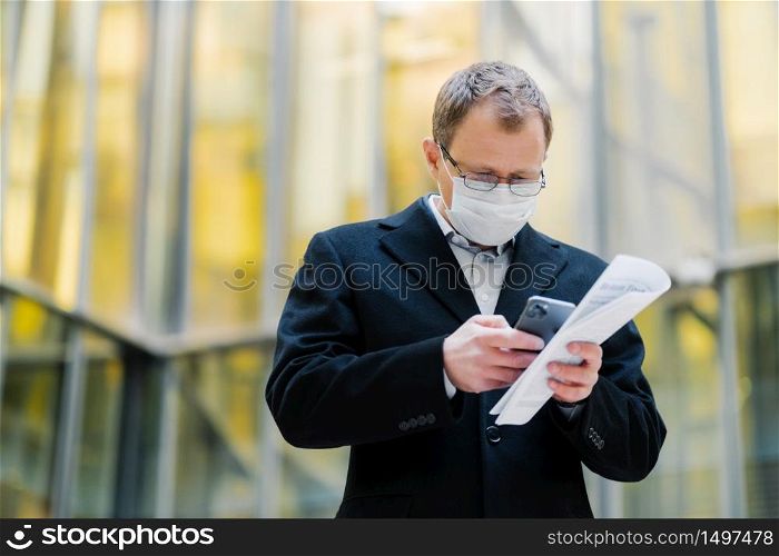 Coronavirus pandemic concept. Photo of male manager focused in smartphone device, sends text messages, wears spectacles and medical mask, holds papers, walks at street during quarantine, pandemic