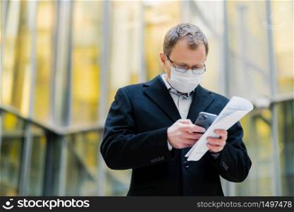 Coronavirus pandemic concept. Photo of male manager focused in smartphone device, sends text messages, wears spectacles and medical mask, holds papers, walks at street during quarantine, pandemic