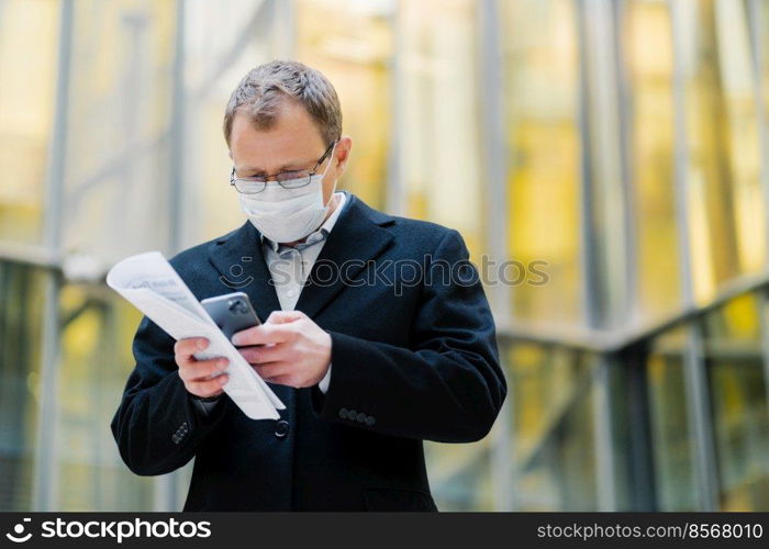 Coronavirus pandemic concept. Photo of male manager focused in smartphone device, sends text messages, wears spectacles and medical mask, holds papers, walks at street during quarantine, pandemic