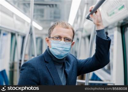 Coronavirus prevention. Man office worker wears transparent glasses, formal outfit, medical mask,  ares about safety, poses in metro train, travels in public transport during Covid-19 outbreak.