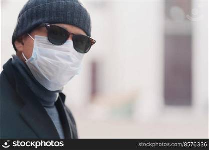 Coronavirus, quarantine and pandemic disease concept. Sideways shot of European man wears shades and disposable medical mask, poses outdoor against blurred background. Health care. Horizontal shot.