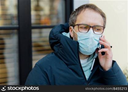 Coronavirus quarantine. Sad puzzled man in panic because of epidemic disease, covers face with medical mask, wears spectacles, keeps mobile phone near ear, consults doctor on distance, stands outdoor