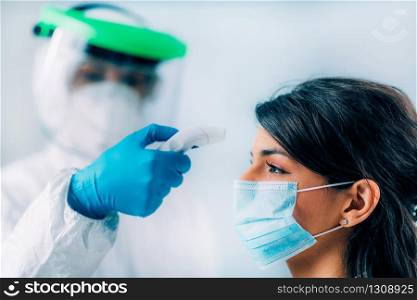 Coronavirus Screening. Medical worker in protective suit measuring body temperature with contactless body thermometer, young woman wearing protective mask. Coronavirus Screening. Medical worker in protective suit checking body temperature with contactless body thermometer, young woman wearing protective mask