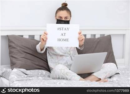 Coronavirus. Young woman in protection mask with laptop holding paper with Stay Home on bed. isolated to prevent infection of pandemic, epidemic Covid-19. Quarantine at home. selective focus. Coronavirus. Young woman in protection mask with laptop holding paper with Stay Home on bed. isolated to prevent infection of pandemic, epidemic Covid-19. Quarantine at home. selective focus.