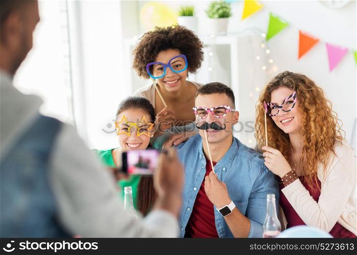 corporate, celebration and holidays concept - man taking picture of happy friends or team with party accessories having fun at office. friends or team photographing at office party