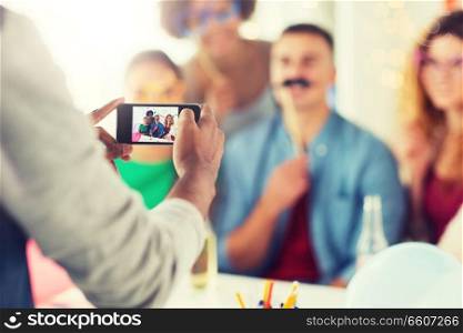 corporate, celebration and holidays concept - man with smartphone taking picture of happy friends or coworkers with party accessories having fun at office. friends or coworkers photographing at office party