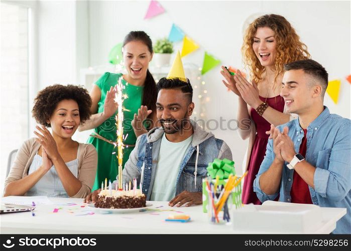 corporate, celebration and people concept - happy team with firework on birthday cake and non-alcoholic drinks greeting colleague at office party. team greeting colleague at office birthday party