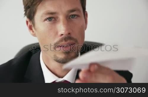 Corporate manager in modern office takes a break and prepares a paper airplane. The bored man thinks of his vacations and leans back on his chair with upset expression. Closeup shot