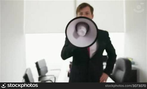 Corporate manager standing with megaphone in modern office. The man screams loud with energetic expression and crosses arms in portrait pose. Steadicam shot