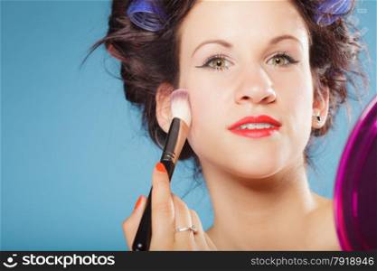 Cosmetic beauty procedures and makeover concept. Woman in hair curlers applying makeup blusher with brush. Girl gets blush on cheekbones, on blue