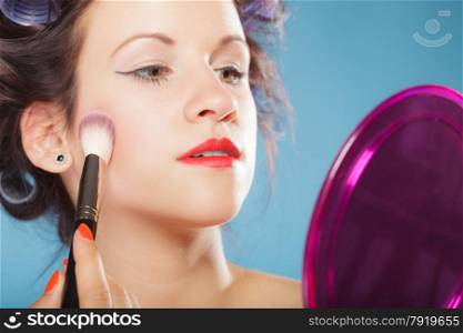 Cosmetic beauty procedures and makeover concept. Woman in hair curlers applying makeup blusher with brush. Girl gets blush on cheekbones, on blue