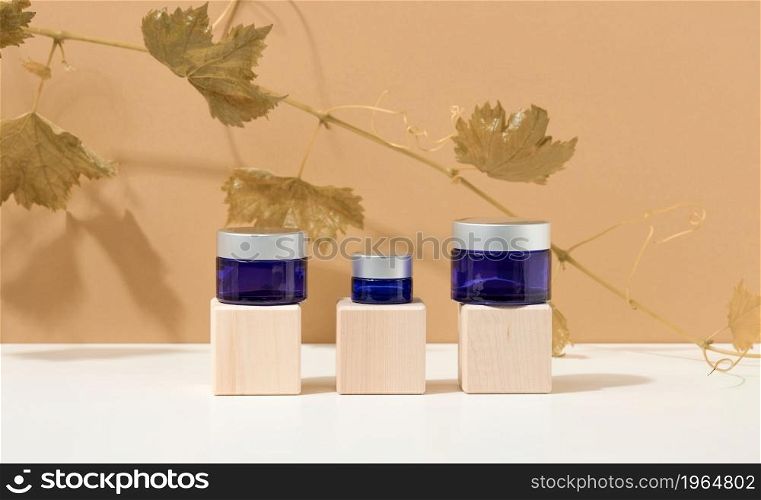 cosmetic products in a blue glass jar with a gray lid on a wooden podium made of cubes, behind a branch of grapes with green leaves. Blank for branding products, moisturizer on beige background