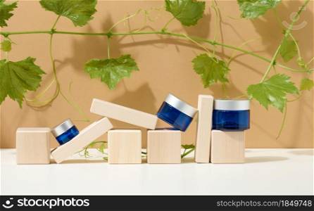 cosmetic products in a blue glass jar with a gray lid stand on a wooden podium made of cubes, behind a branch of grapes with green leaves. Blank for branding products, moisturizer on beige background