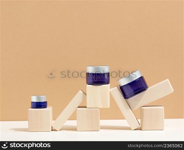 cosmetic products in a blue glass jar with a gray lid stand on a wooden podium made of cubes. Blank for branding products, moisturizer on beige background