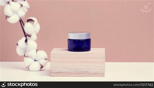 cosmetic products in a blue glass jar with a gray lid stand on a wooden podium made of cubes. Blank for branding products, moisturizer on beige background