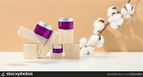 cosmetic products in a purple glass jar with a gray lid on a wooden podium made of cubes, behind a branch of grapes with green leaves. Blank for branding products, moisturizer on beige background