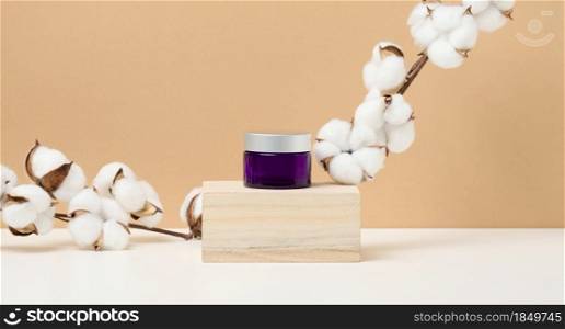 cosmetic products in a purple glass jar with a gray lid stand on a wooden podium made of cubes. Blank for branding products, moisturizer on beige background