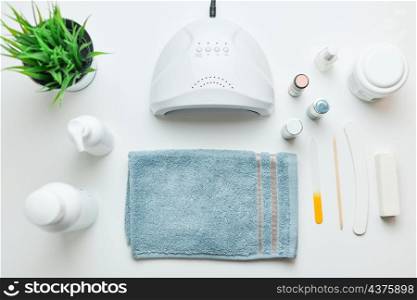 Cosmetic products set, nail hybrid gels, UV lamp, green leaves, blue towel on white table. Spa, manicure, skin care concept. Flat lay, overhead view
