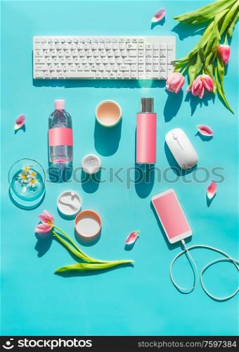 Cosmetic products with mock up on turquoise desktop. PC keyboard , smartphone with blank screen and wire and pink tulips flowers in sunlight. Top view. Flat lay. Springtime. Blog layout. Home office