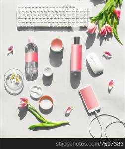 Cosmetic products with mock up on white desktop. PC keyboard , smartphone with blank screen and wire and pink tulips flowers in sunlight. Top view. Flat lay. Springtime. Beauty blog layout.