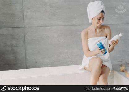 Cosmetics choosing. Lady is holding a flacons with cosmetics. Micellar water or face toner bottles. Gorgeous caucasian woman wrapped in towel after bathing. Young woman takes shower at home.. Cosmetics choosing. Lady is holding a flacons with cosmetics. Micellar water or face toner bottles.