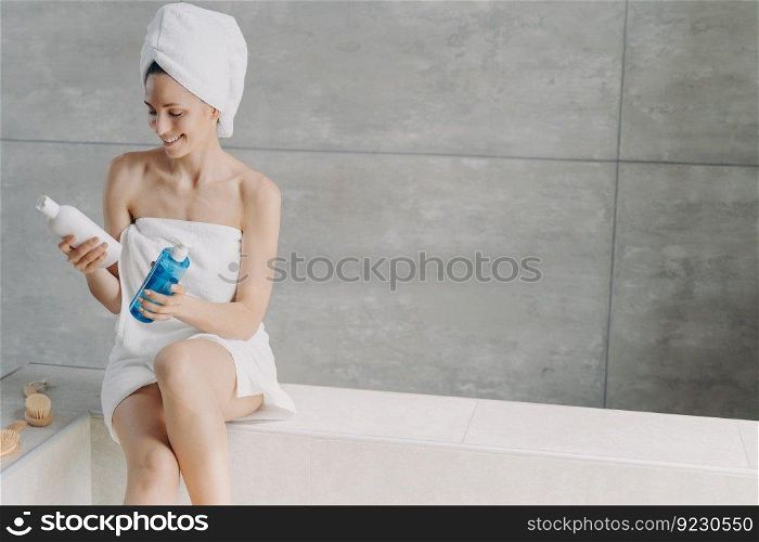 Cosmetics choosing. Lady is holding a flacons with cosmetics. Micellar water or face toner bottles. Gorgeous caucasian woman wrapped in towel after bathing. Young woman takes shower at home.. Cosmetics choosing. Lady is holding a flacons with cosmetics. Micellar water or face toner bottles.
