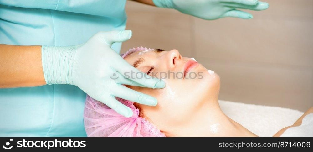 Cosmetologist with gloved hands applies a moisturizing mask with peeling cream on the female face. Facial cosmetology treatment. Procedures for facial care. Cosmetologist with gloved hands applies a moisturizing mask with peeling cream on the female face. Facial cosmetology treatment. Procedures for facial care.