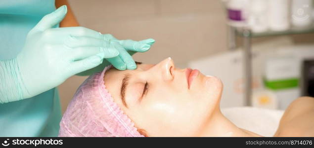 Cosmetologist with gloved hands applies a moisturizing mask with peeling cream on the female face. Facial cosmetology treatment. Procedures for facial care. Cosmetologist with gloved hands applies a moisturizing mask with peeling cream on the female face. Facial cosmetology treatment. Procedures for facial care.