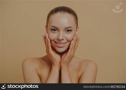 Cosmetology and dermatology. Headshot of beautiful woman touching cheeks, enjoying her soft and smooth skin, standing half-naked over beige background. Face of smiling european lady without make-up. Young beautiful woman with bare shoulders touching cheeks, enjoying her soft and smooth skin