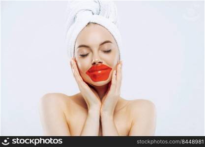Cosmetology and skin care concept. Pretty woman with calm expression, closes eyes, applies lips patches, wears natural makeup, wrapped towel on head, stands bare shoulders indoor, white background