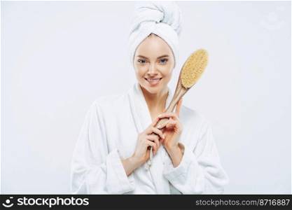 Cosmetology, peeling, spa and grooming concept. Pleased young European woman with healthy skin, gentle smile, holds massage brush for body and feet, cares about herself, dressed in bath robe