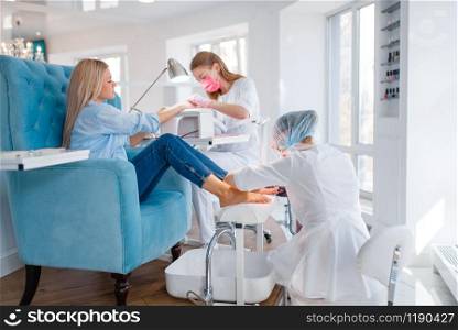 Cosmetology salon, manicure and pedicure procedure. Female client in beauty shop, woman at the cosmetologist, two masters works with fingernails and toenails. Cosmetology salon, manicure and pedicure procedure