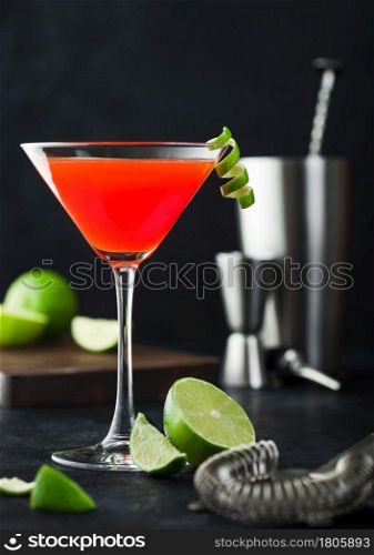 Cosmopolitan cocktail in classic crystal glass with lime peel and fresh limes with strainer, jigger and shaker on black table background.